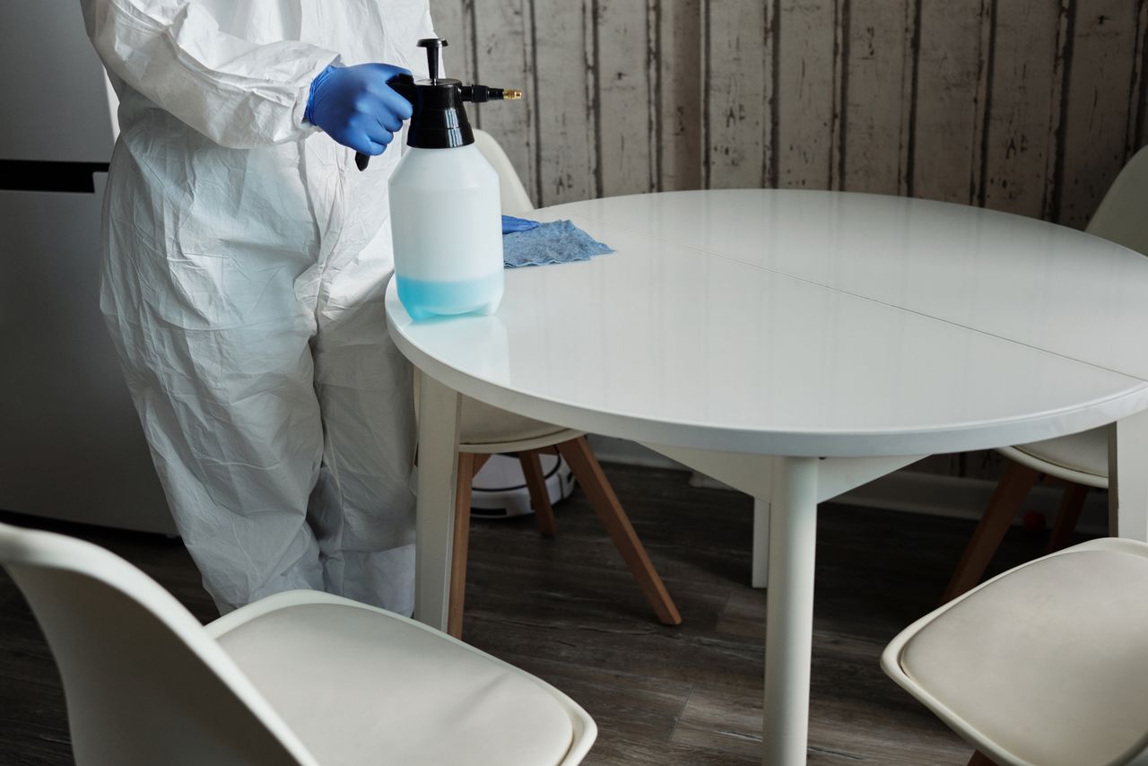 team-04 A person wearing a protective suit is disinfecting a white indoor table with a sprayer.